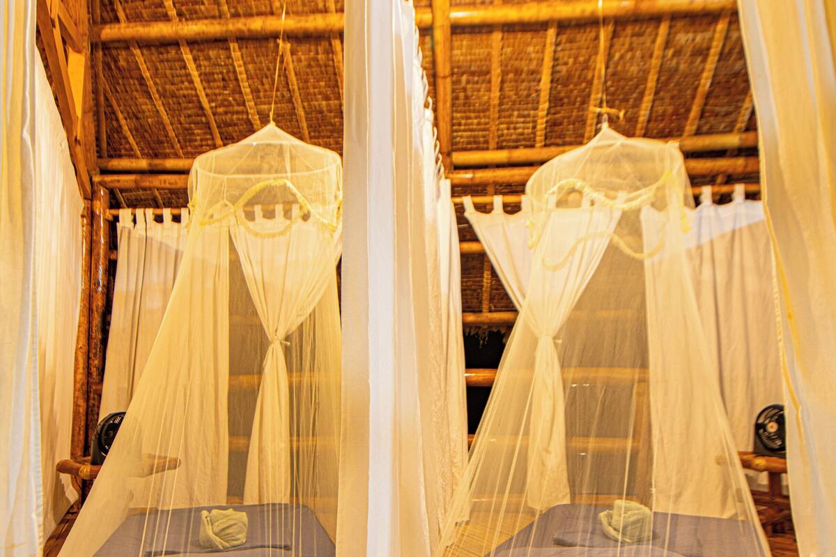 A room with two beds, each with a canopy. The room has a wooden floor and a thatched roof.
