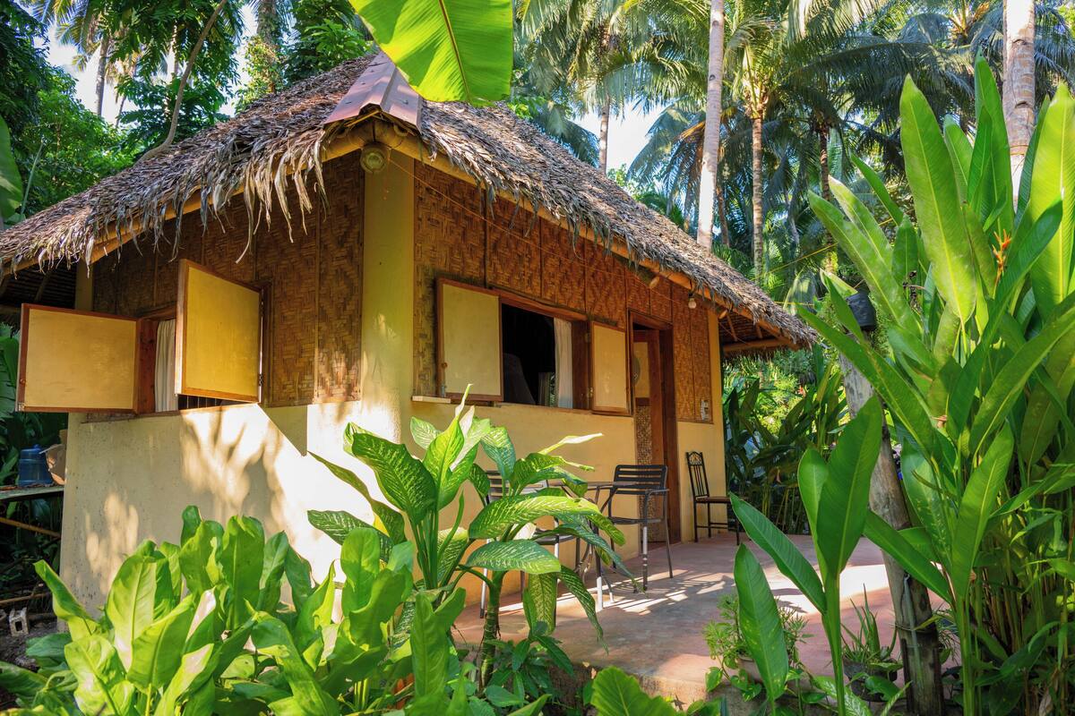 A rustic thatched-roof hut with a veranda, surrounded by lush tropical vegetation.