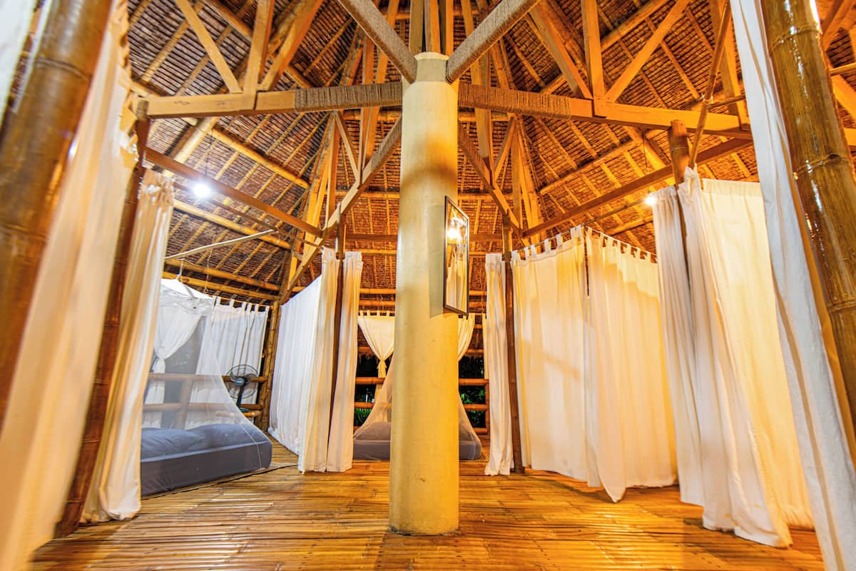 A room with a wooden ceiling and a large pillar in the center. There is a bed with white curtains and a mattress.