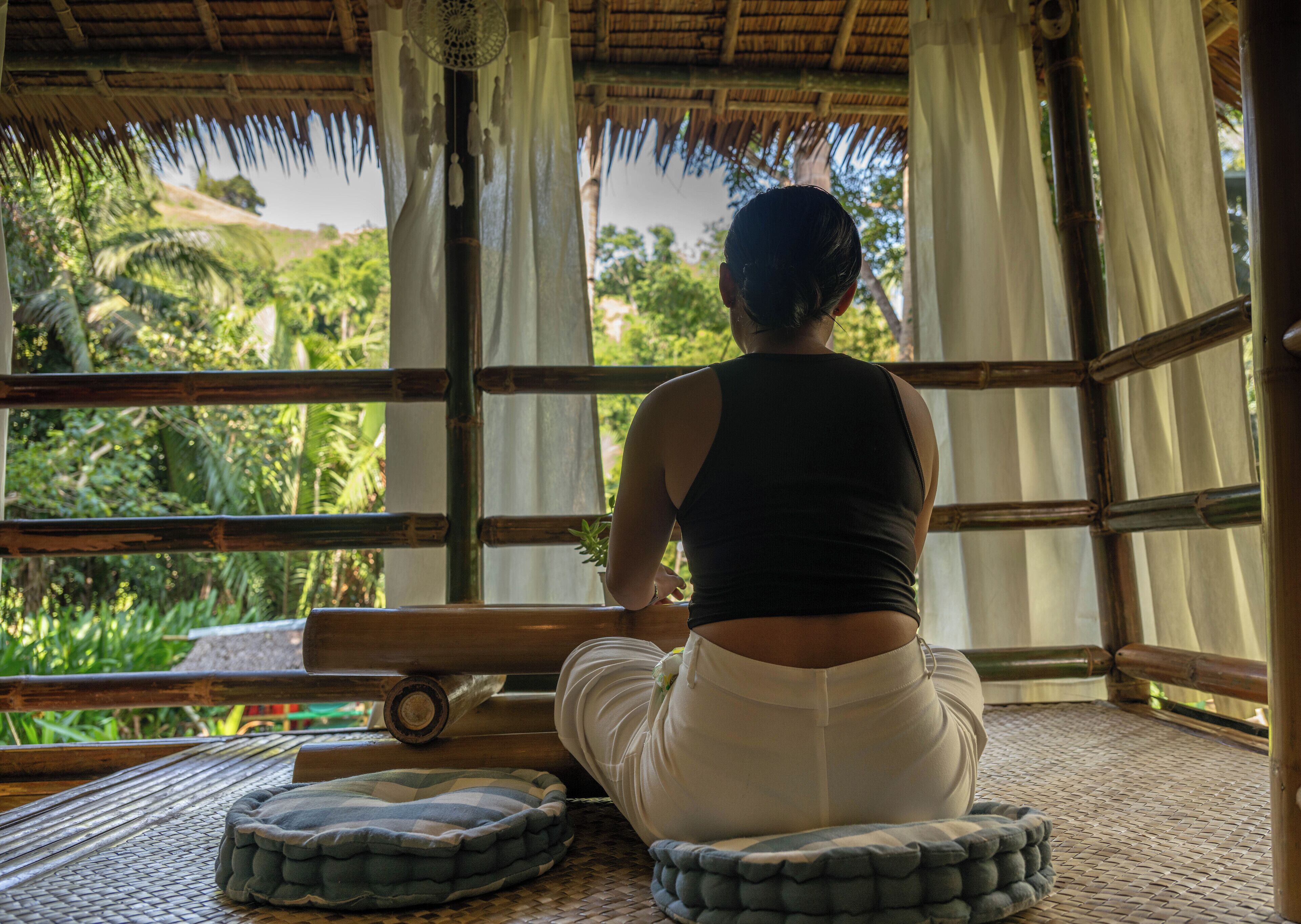 Una persona sentada sobre un tapete tejido dentro de una estructura de bambú con vistas a exuberante follaje.