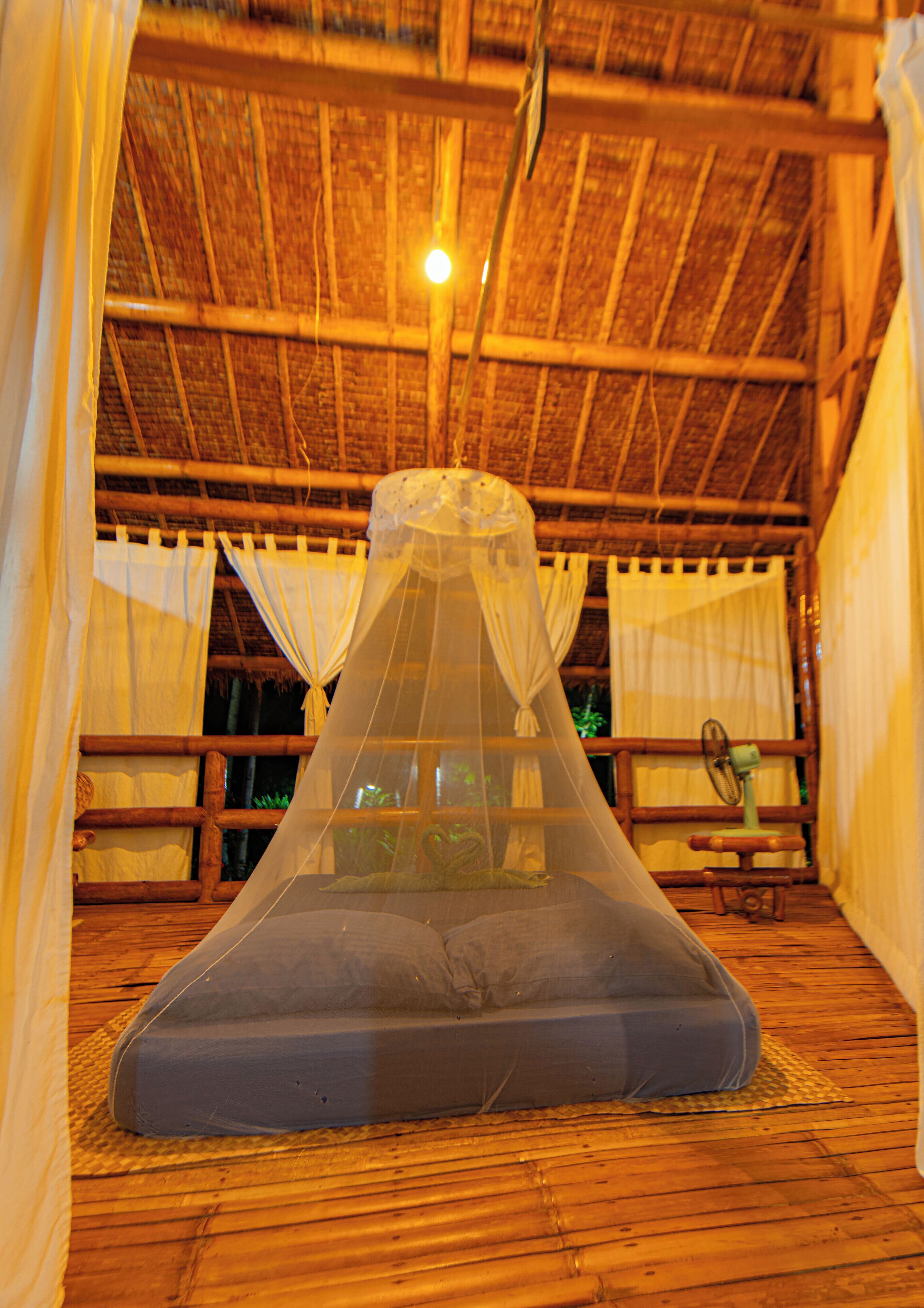 Una habitación con techo de paja, una cama con mosquitero y una mesita con ventilador.
