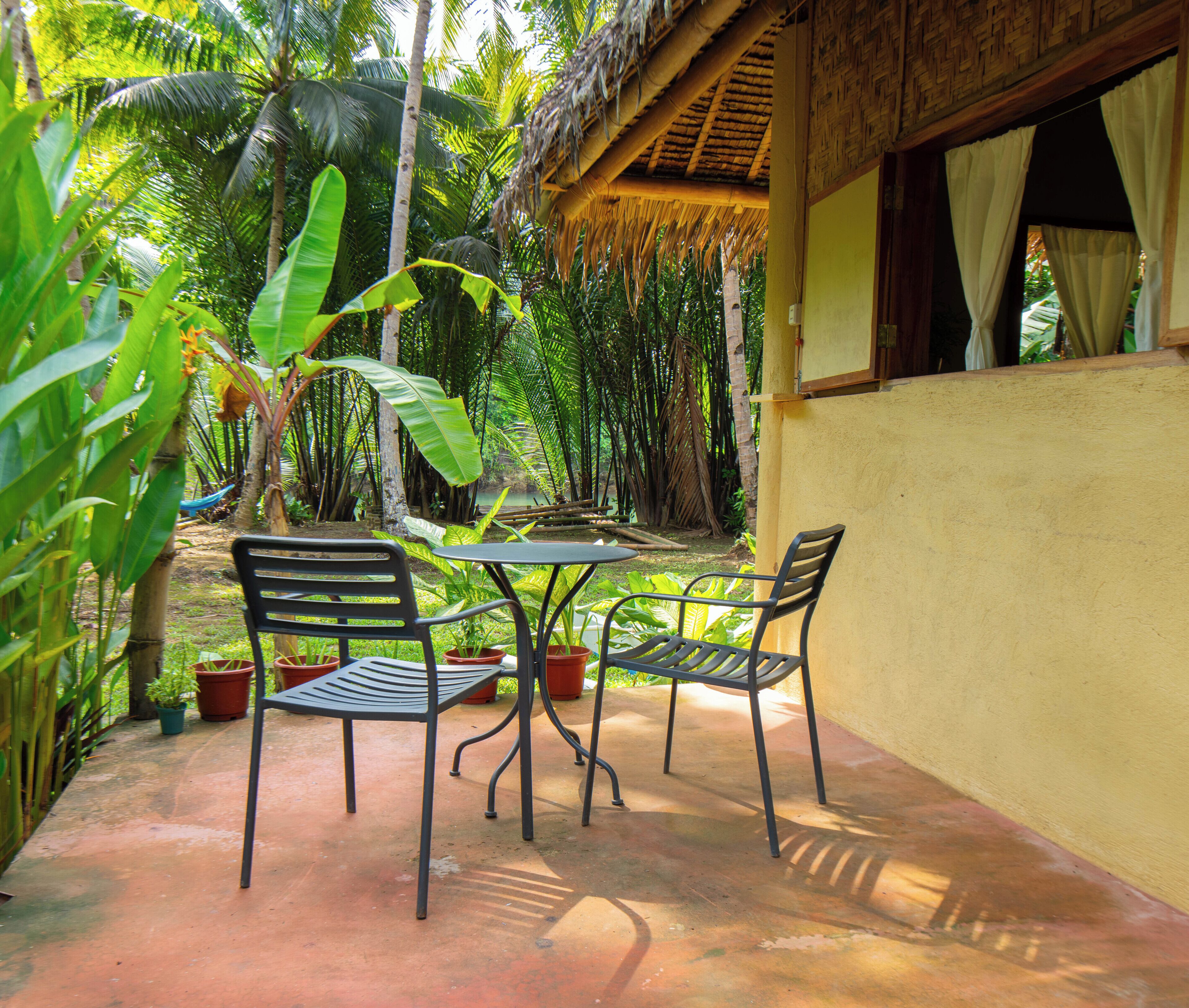 An outdoor seating area with two metal chairs and a table, surrounded by lush greenery and a thatched roof structure.