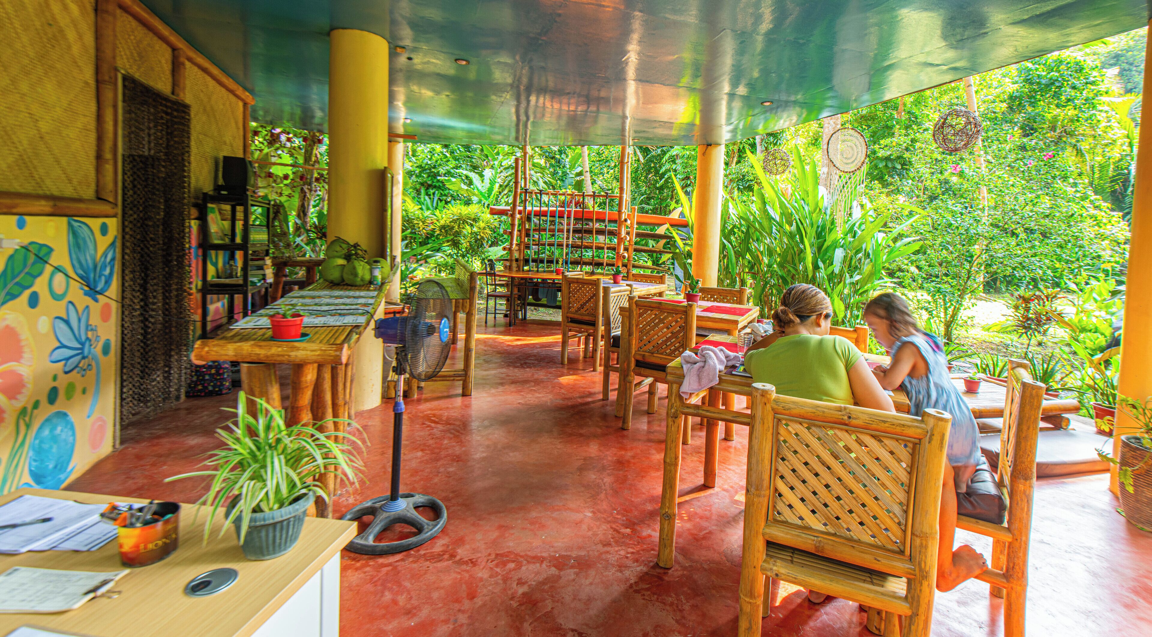 Un área de comedor al aire libre cubierta, con mobiliario de madera y plantas en macetas.
