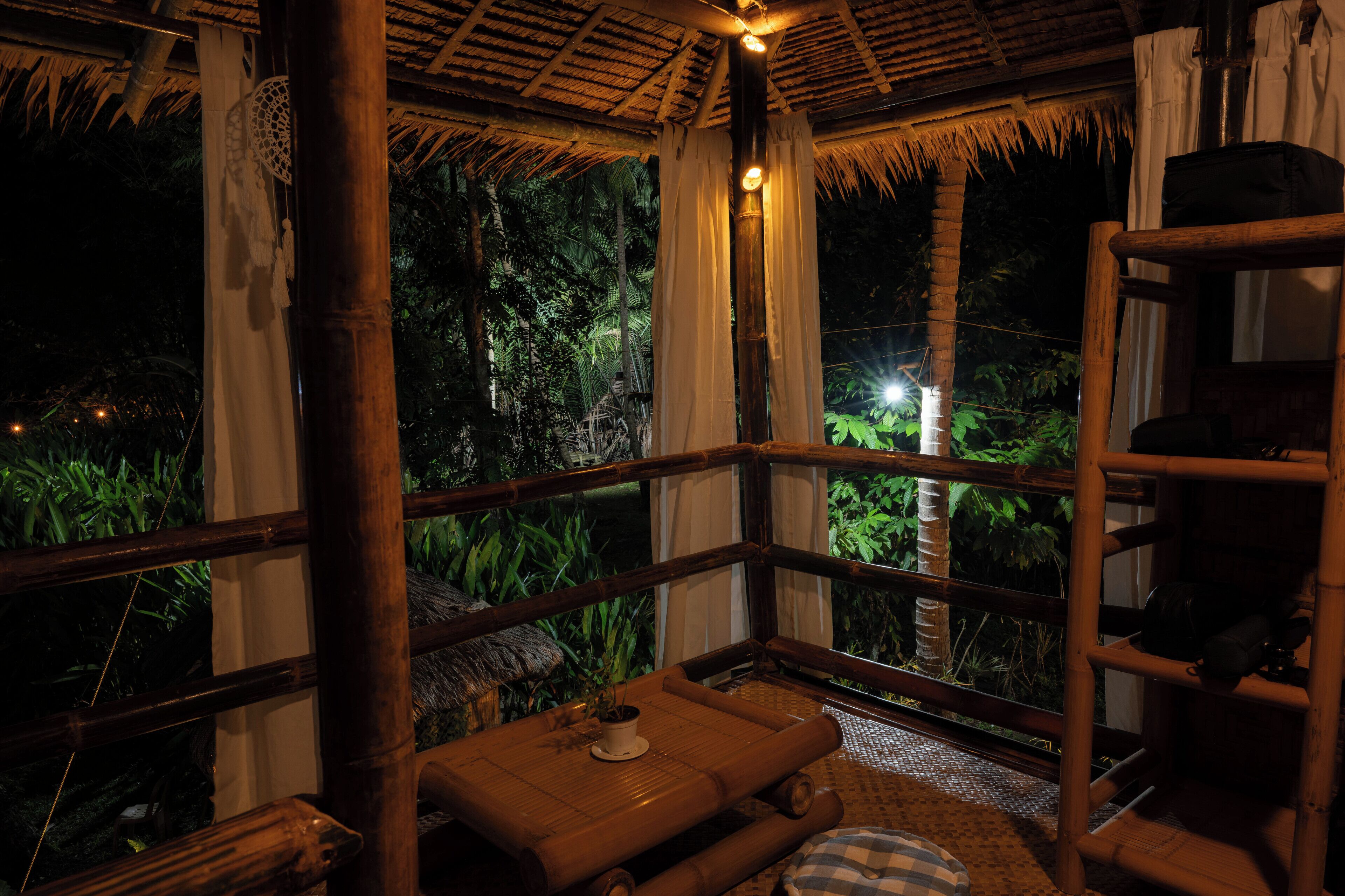 Una habitación con estructura de bambú y techo de paja, un banco de madera y vistas a exuberante vegetación.