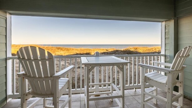 A covered balcony with two Adirondack chairs and a small table, overlooking a beach with golden sand and a clear sky. - Peaceful Oceanfront OBX Getaway (Kill Devil Hills)