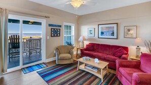 A living room with a red sofa, a beige armchair, a coffee table, and a ceiling fan. There is a sliding glass door leading to a balcony with outdoor furniture. - Peaceful Oceanfront OBX Getaway (Kill Devil Hills)