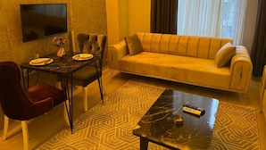 A living room with a sofa, a dining table with chairs, a TV mounted on the wall, and a patterned rug. - Vast Apart Otel (Izmir)