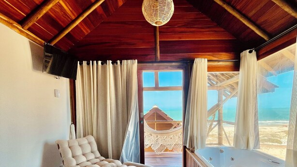 Una habitación acogedora con techo de madera, un sofá cómodo, una butaca blanca acolchada, una bañera y vistas a la playa a través de un ventanal grande.