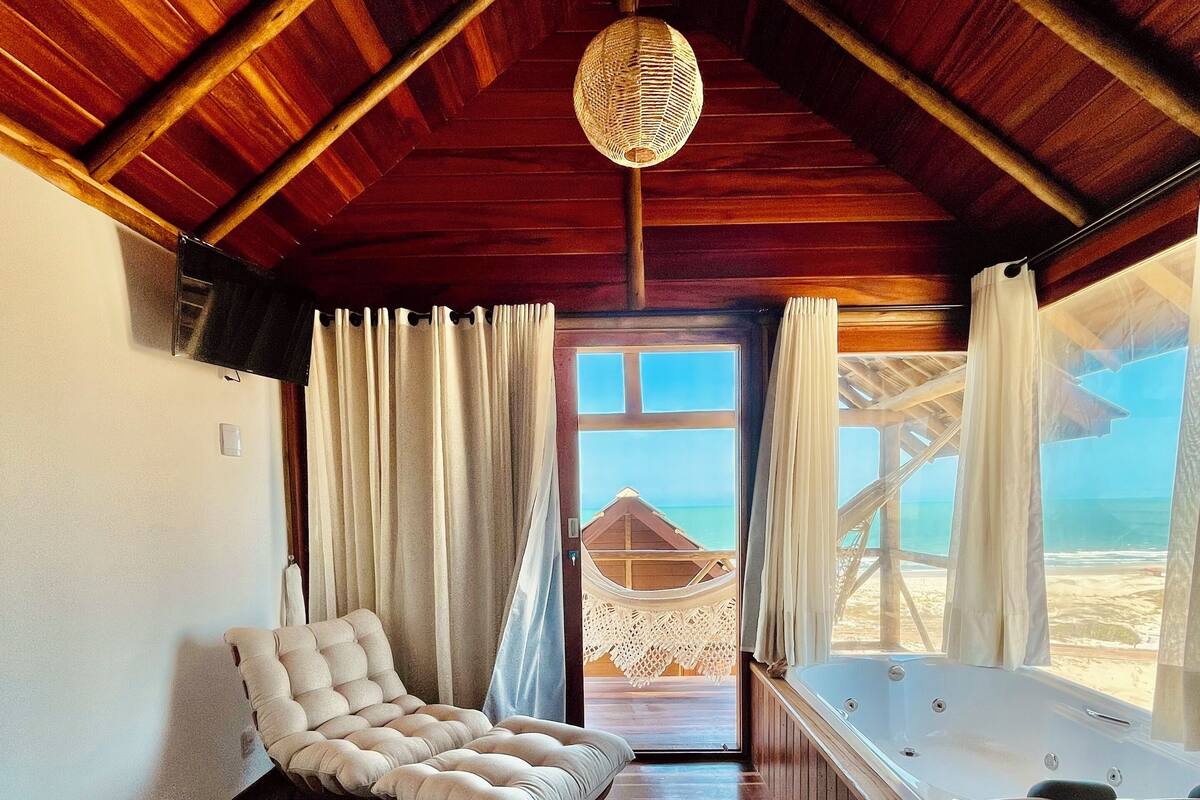 Una habitación acogedora con techo de madera, un sofá cómodo, una butaca blanca acolchada, una bañera y vistas a la playa a través de un ventanal grande.