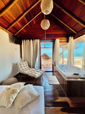 A cozy room with a wooden ceiling, a comfortable sofa, a white cushioned chair, a bathtub, and a view of the beach through a large window. - Vila Jung Bangalôs de Charme (Beberibe)