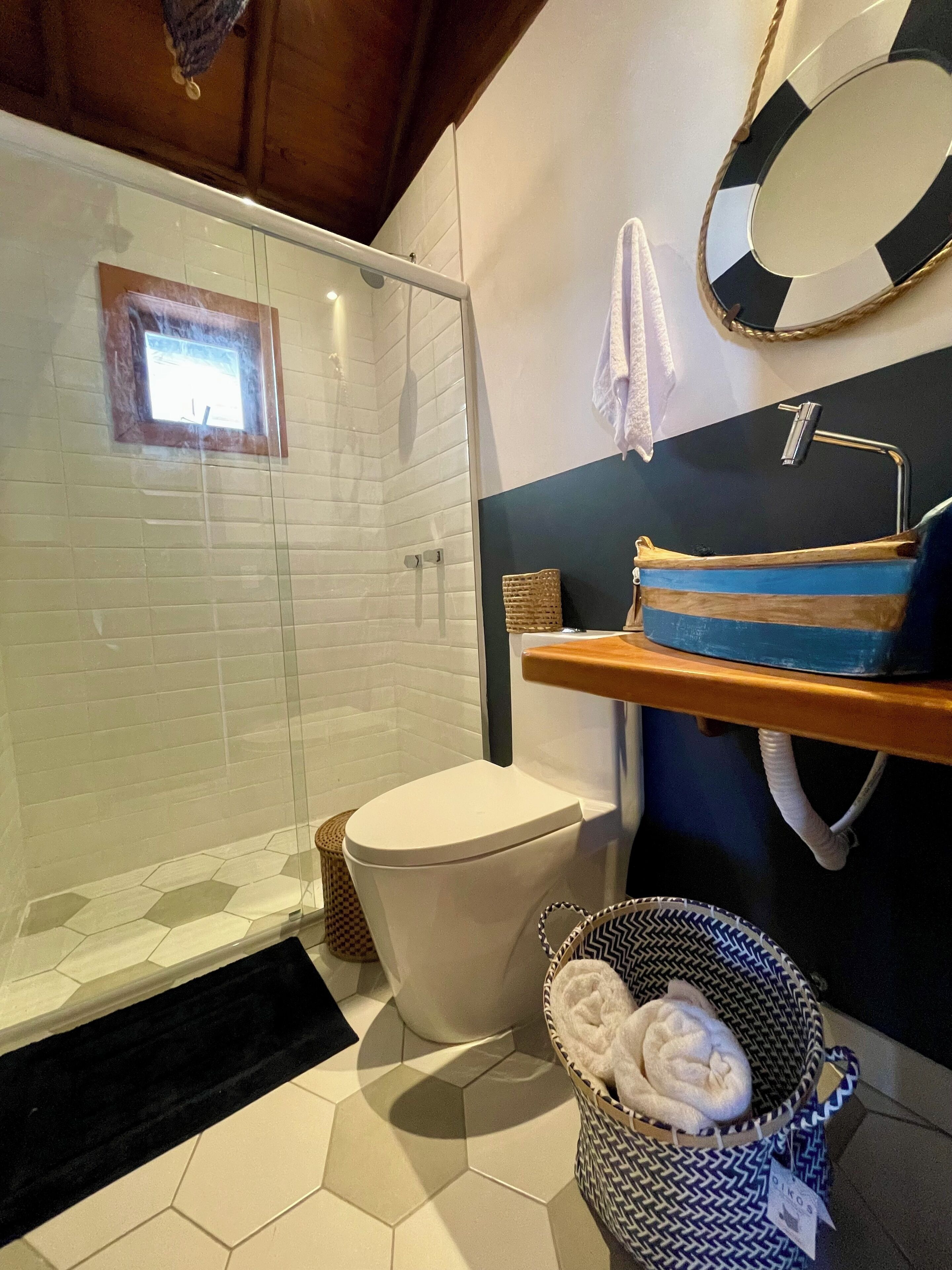 Un baño con ducha de mampara de vidrio, inodoro blanco, cesta azul y blanca con toallas, estante de madera con una toalla azul, y un espejo redondo con marco blanco y negro.