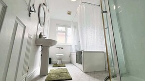 A bathroom with a white sink, toilet, and bathtub, a shower curtain, and a window. - Spacious 4-Bed House I Free Parking & Fast Wi-Fi (Dagenham)