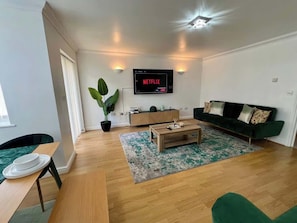 En stue med trægulv, grøn sofa, sofabord, fjernsyn ophængt på væggen og en potteplante.