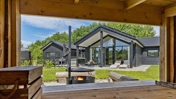 Une terrasse extérieure moderne, dotée d’un foyer, de meubles en bois et offrant une vue sur une maison contemporaine avec de grandes fenêtres et une pelouse.