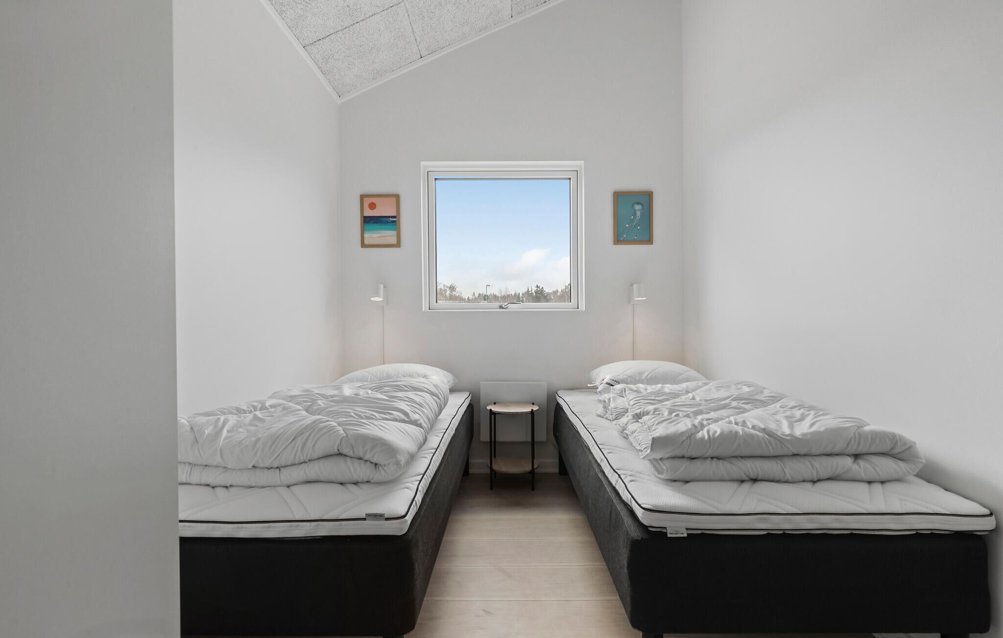 A small, modern bedroom with two single beds, a small table, and a window with a view of trees and a clear sky.