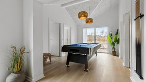 Une table de billard dans une pièce au plancher en bois, aux murs blancs et au plafond haut avec des poutres apparentes.