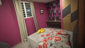 Un dormitorio con cama, una mesita, una silla, una ventana con cortinas y un cuadro en la pared.