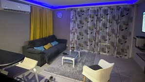 Una sala de estar con sofá, mesa de centro y sillas. Hay un televisor a la derecha y un equipo de aire acondicionado a la izquierda.