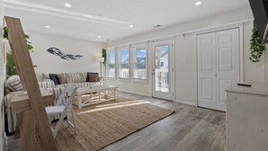 Ein modernes Wohnzimmer mit Sofa, Couchtisch und Teppich. Es gibt Fenster mit Jalousien, eine Tür und eine Pflanze an der Wand.