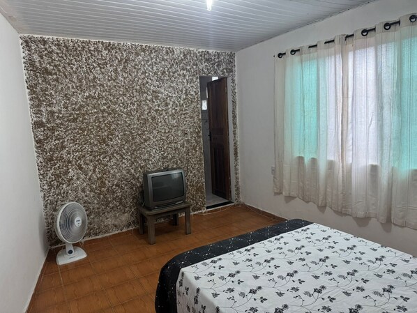Ein Zimmer mit einem Bett, einem Fernseher, einem Ventilator und einem Fenster mit Vorhängen.