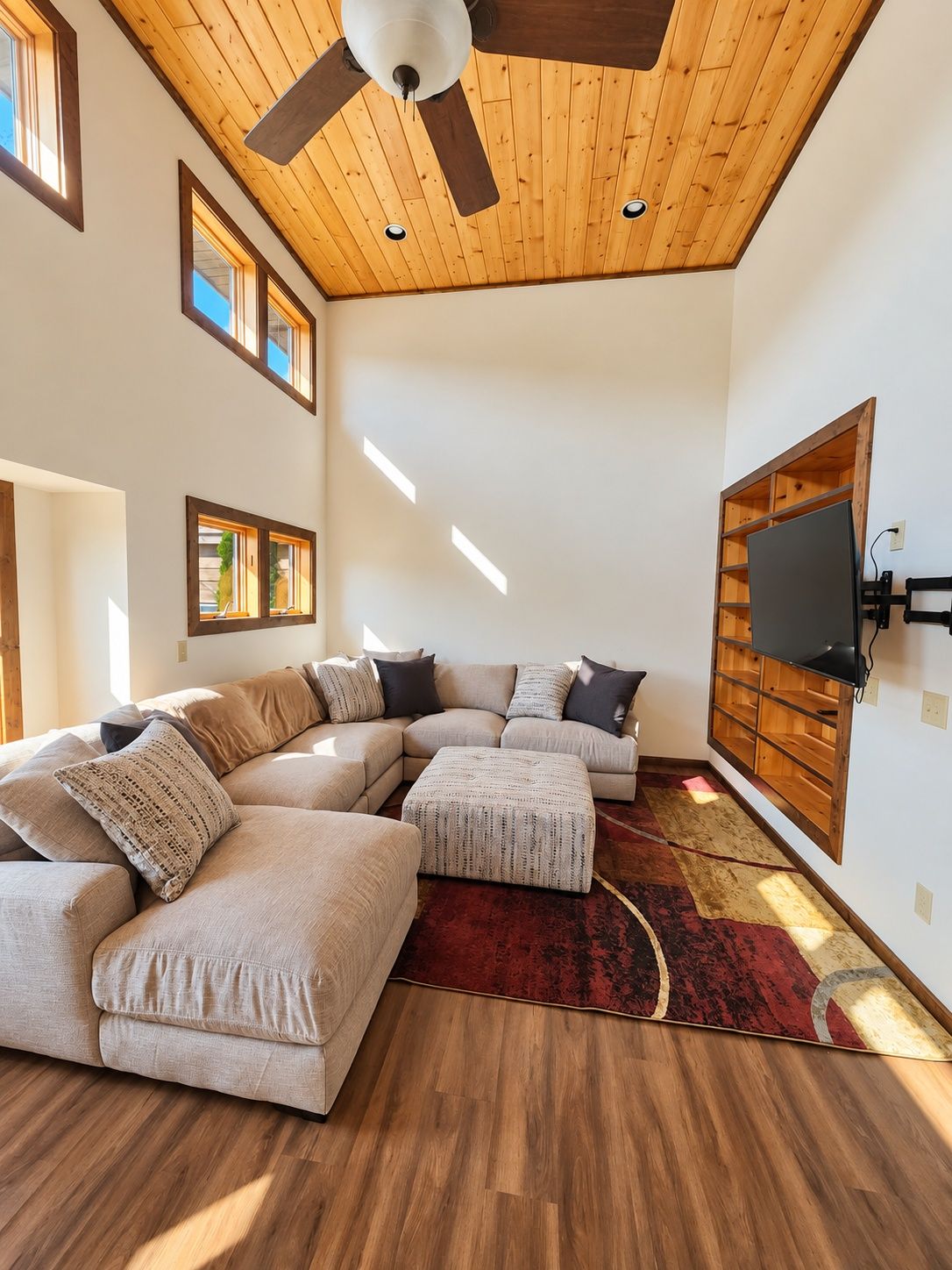 Un salon moderne avec un plafond en bois, un canapé d’angle, un téléviseur à écran plat fixé à un mur en bois et un tapis rouge à motifs.
