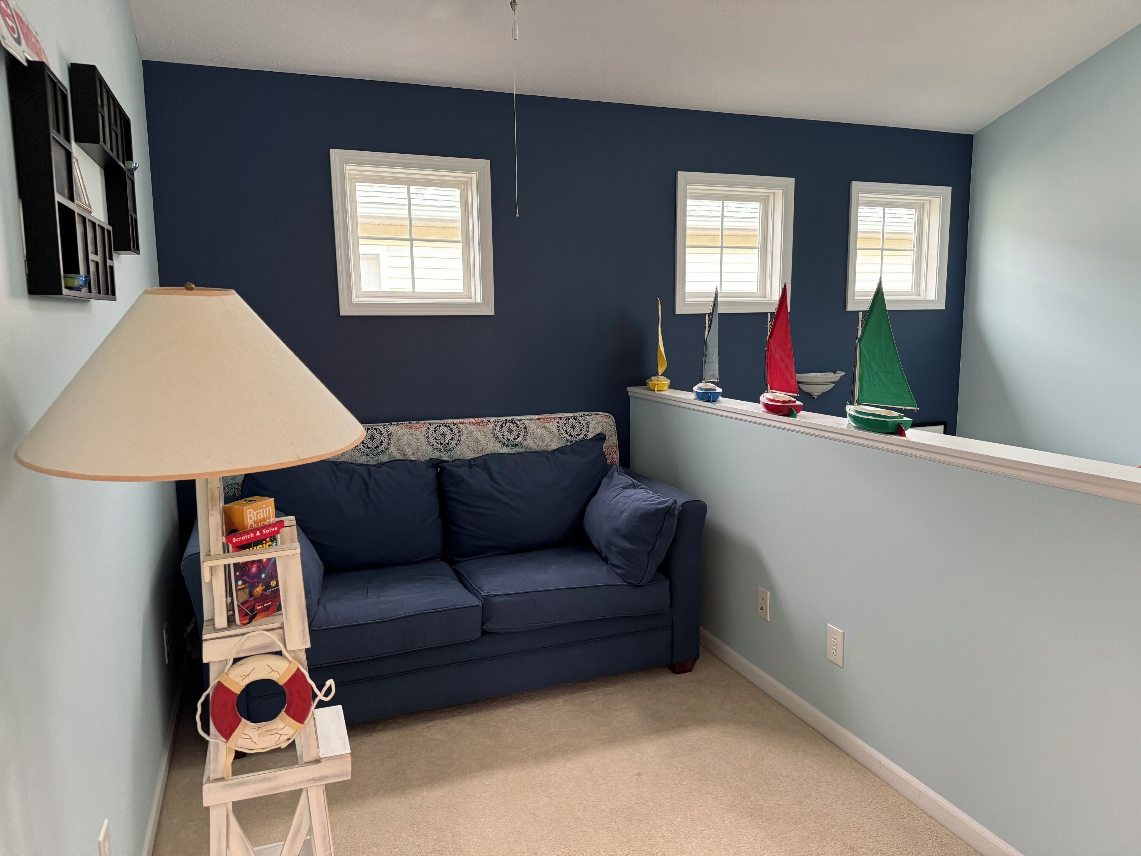 A room with a dark blue wall, a blue sofa, a small shelf with model sailboats, a lamp, and a bookshelf.