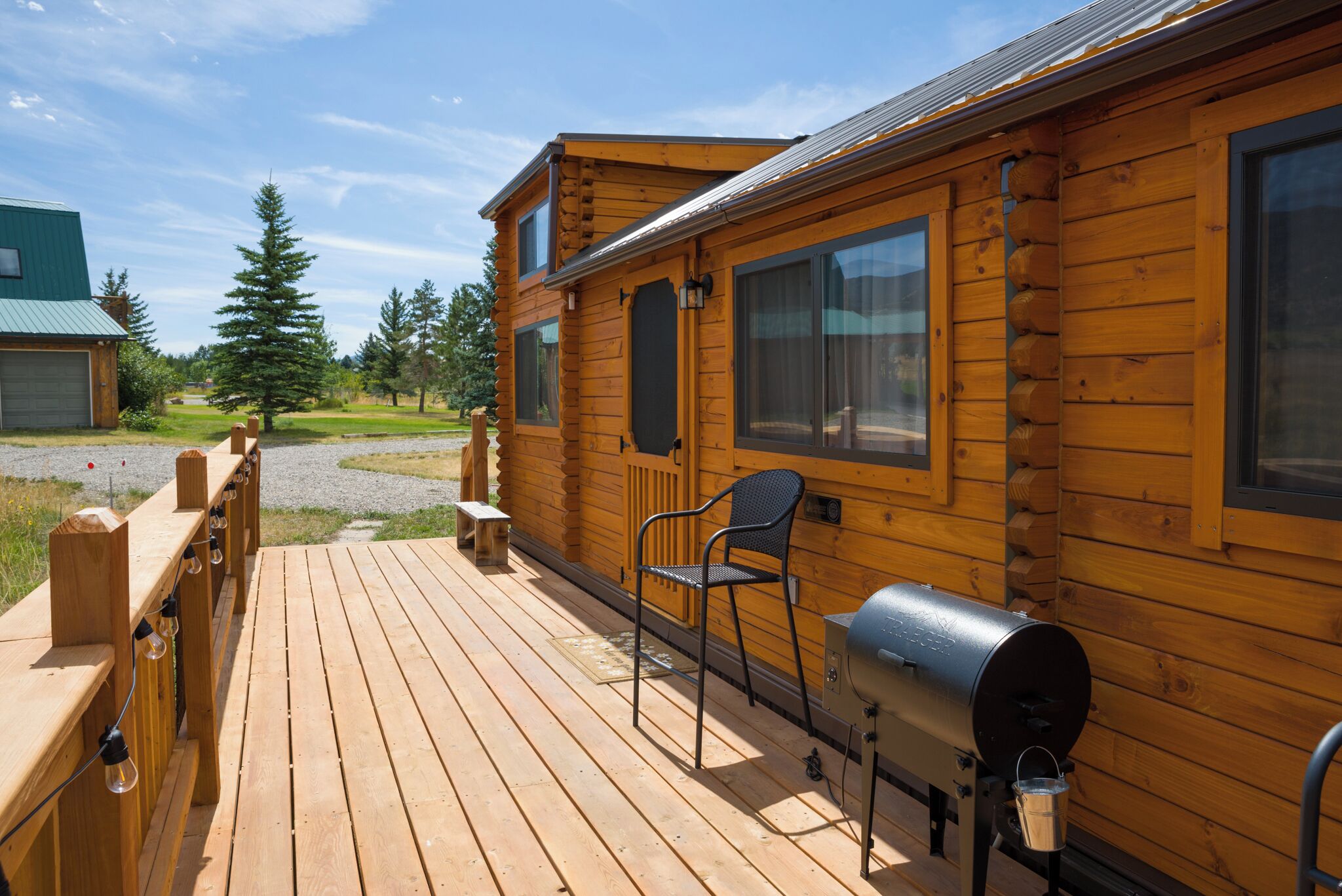 A wooden deck with a grill and a chair, a wooden cabin with large windows, and a view of the surrounding landscape.