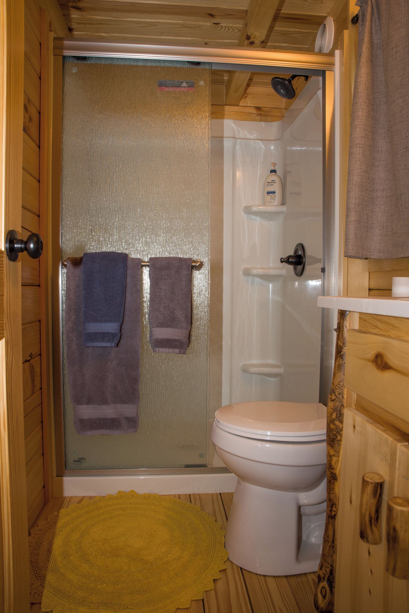 A wooden cabin bathroom with a glass shower enclosure, a toilet, and a yellow bath mat.