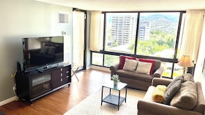 A living room with a flat-screen TV, a sofa, a coffee table, and a large window with a view of buildings and greenery.