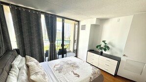 A bedroom with a bed, a dresser, a plant, and a view of the city through a window.