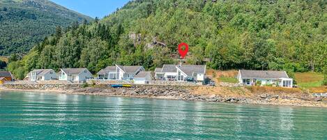 A coastal property with multiple houses along a rocky shoreline, a clear view of the sea, and a forested hillside in the background.