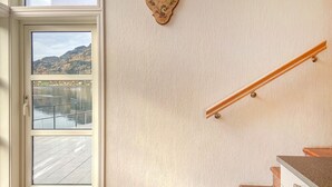 Ein Zimmer mit großem Fenster mit Blick auf einen See und Hügel, eine Holzhandläufe an einer Treppe und eine dekorative Wanduhr.