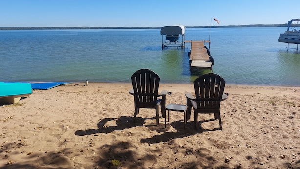 A lakeside setting with a wooden dock extending into the water, two Adirondack chairs facing the water, and a covered boat at the dock's end.