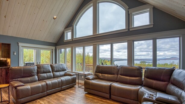A spacious living room with a large sectional sofa, wooden flooring, and large windows offering a view of a lake and mountains. - Lodges #72 - Rangeley Lake Overlook - ATV, Snowmobile, and close to Saddleback! (Rangeley)