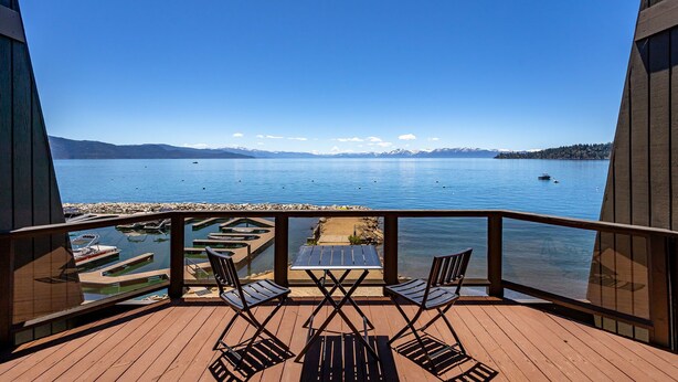 Terraza de madera con dos sillas y una mesa, con vista a un lago tranquilo con barcos y un muelle.