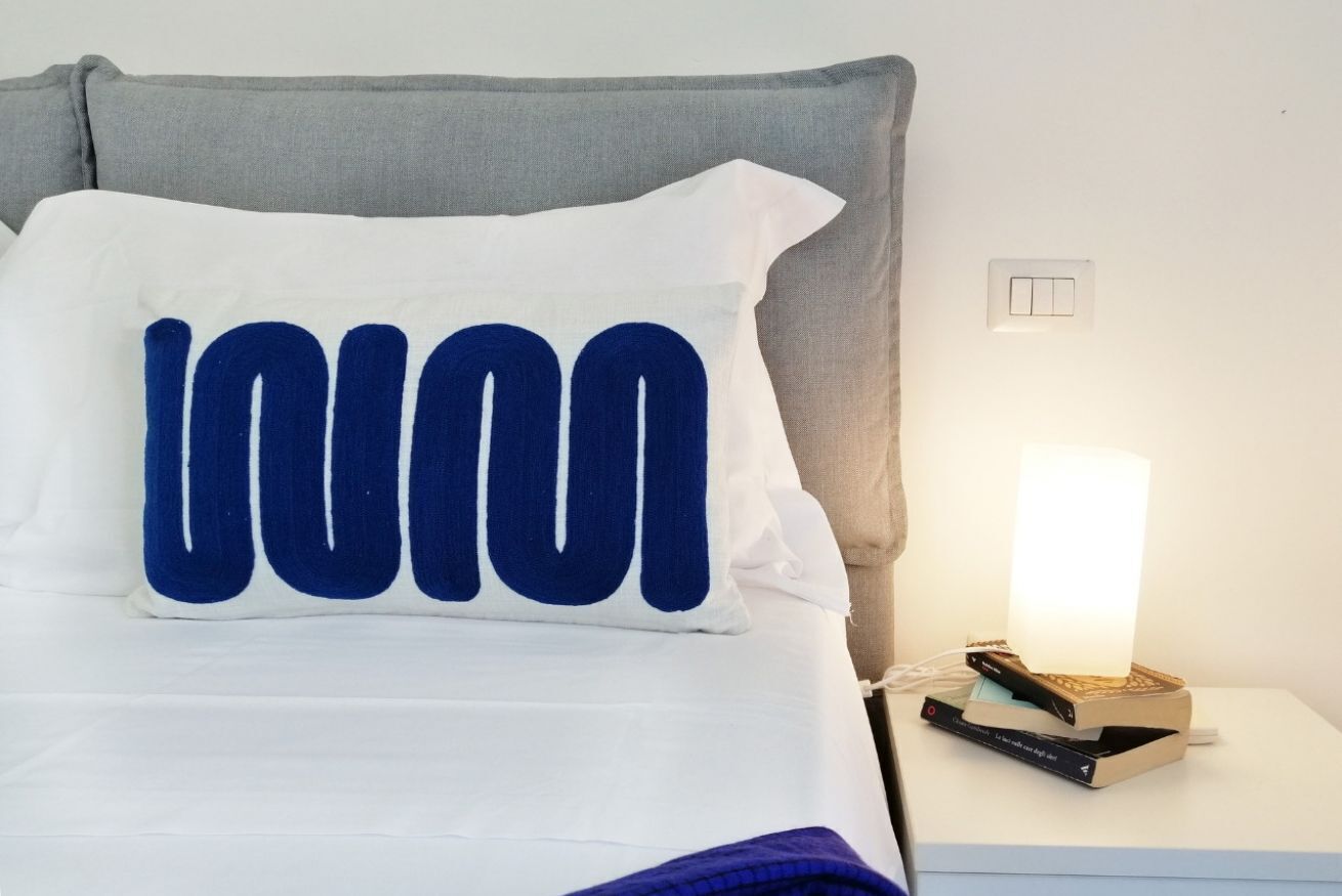 A bed with a white and blue pillow, a gray headboard, a bedside table with a lamp, and a book.