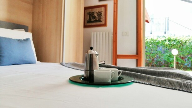 A neatly made bed with a blue pillow, a tray with a coffee pot and cups, and a window with a view of greenery and a building. - Holihome_The Lemon Garden (Tortoreto Lido)