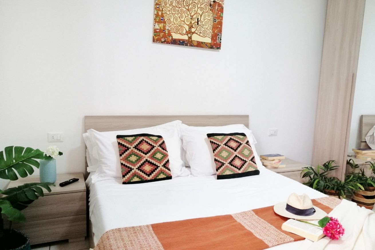 Un lit bien fait, agrémenté d’oreillers à motifs, une table de chevet avec une télécommande, un vase avec une plante et un tableau accroché au mur.