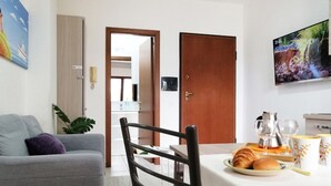A living room with a grey sofa, a dining table with a plate of food, a TV, and a wooden door. - Holihome_Open House FRN (Alba Adriatica)