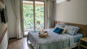 A bedroom with a large bed, a TV mounted on the wall, a balcony with a table and chairs, and a view of greenery outside. - Rio Centro Stay (Rio de Janeiro)