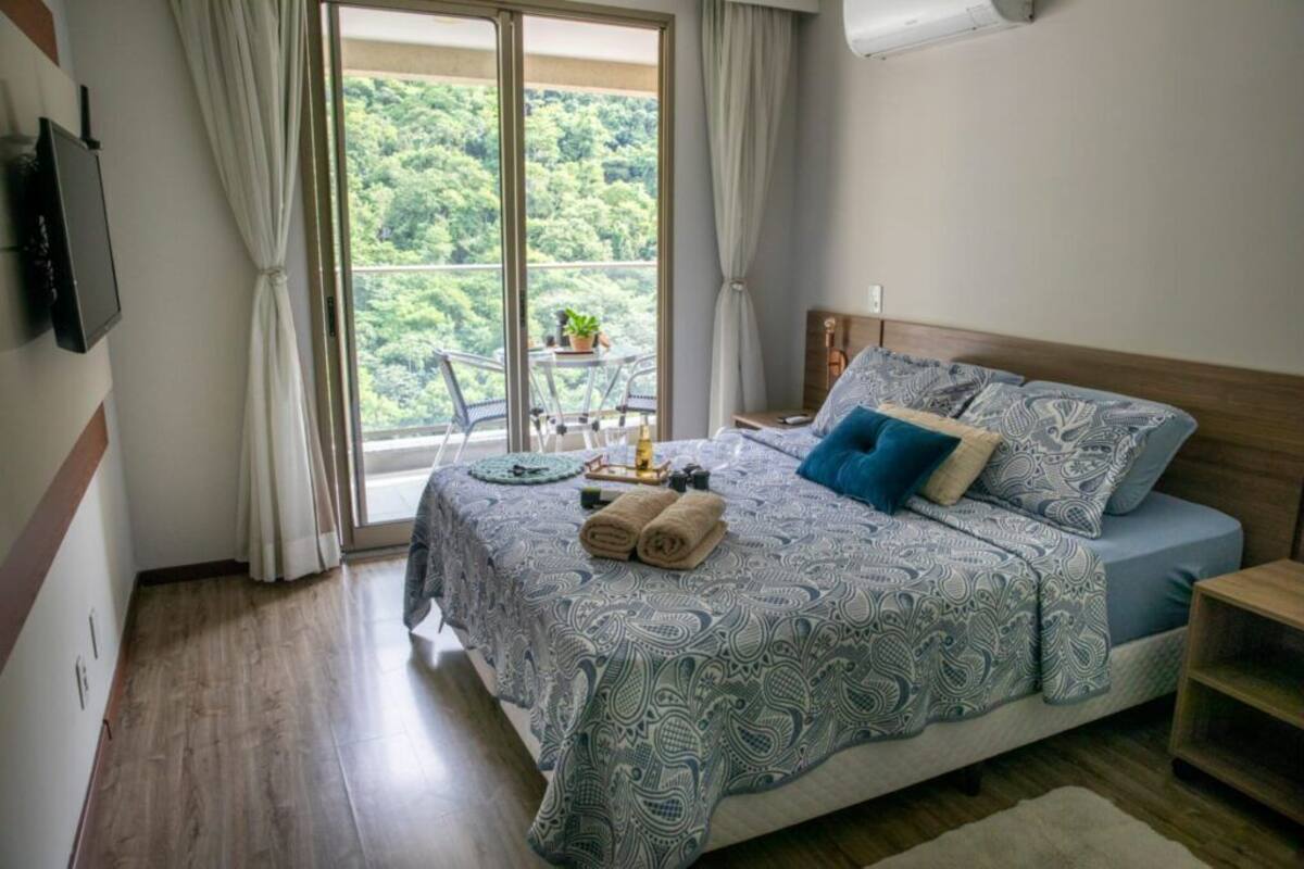 A bedroom with a large bed, a TV mounted on the wall, a balcony with a table and chairs, and a view of greenery outside.