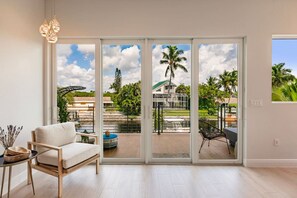 A spacious room with a large sliding glass door leading to a balcony with a view of a tropical landscape, including palm trees and a water feature. - New(2024)Waterfront/Gulf access, Oasis (Bonita Springs)