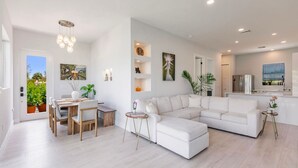En rummelig opholdsstue med hvid sofa, spisebord og stole. Der er et køkkenområde med moderne hårde hvidevarer og en balkon med plantekasser.