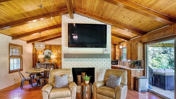 A spacious living room with a fireplace, leather chairs, a dining table, and a large TV mounted on a brick wall. - Fika on The Guadalupe- a charming RIVERFRONT retreat that sleeps 13. (Seguin)