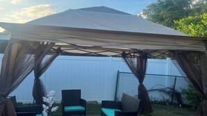 An outdoor patio area with a canopy, two wicker chairs with green cushions, and a small table. - Charming 3-bedroom house in pleasant Port Richey with AC, WiFi (Port Richey)