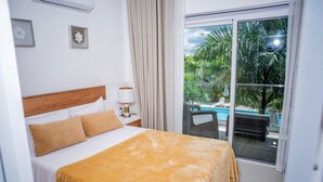 Habitación de hotel con cama, mesita de noche, lámpara y vista a una piscina a través de un ventanal.
