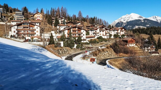 Un village de montagne enneigé, avec des bâtiments à plusieurs étages, une route sinueuse et un ciel bleu dégagé.