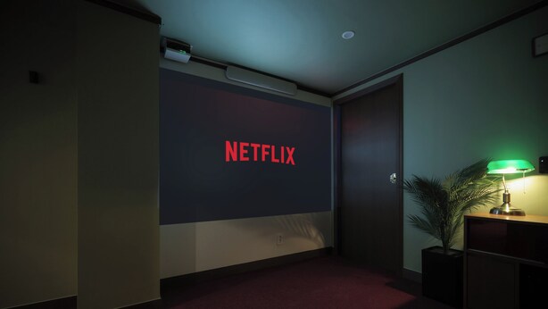 Ein Zimmer mit einer dunklen Wand, dem Netflix-Logo, einem roten Teppich, einer grünen Lampe und einer Pflanze.