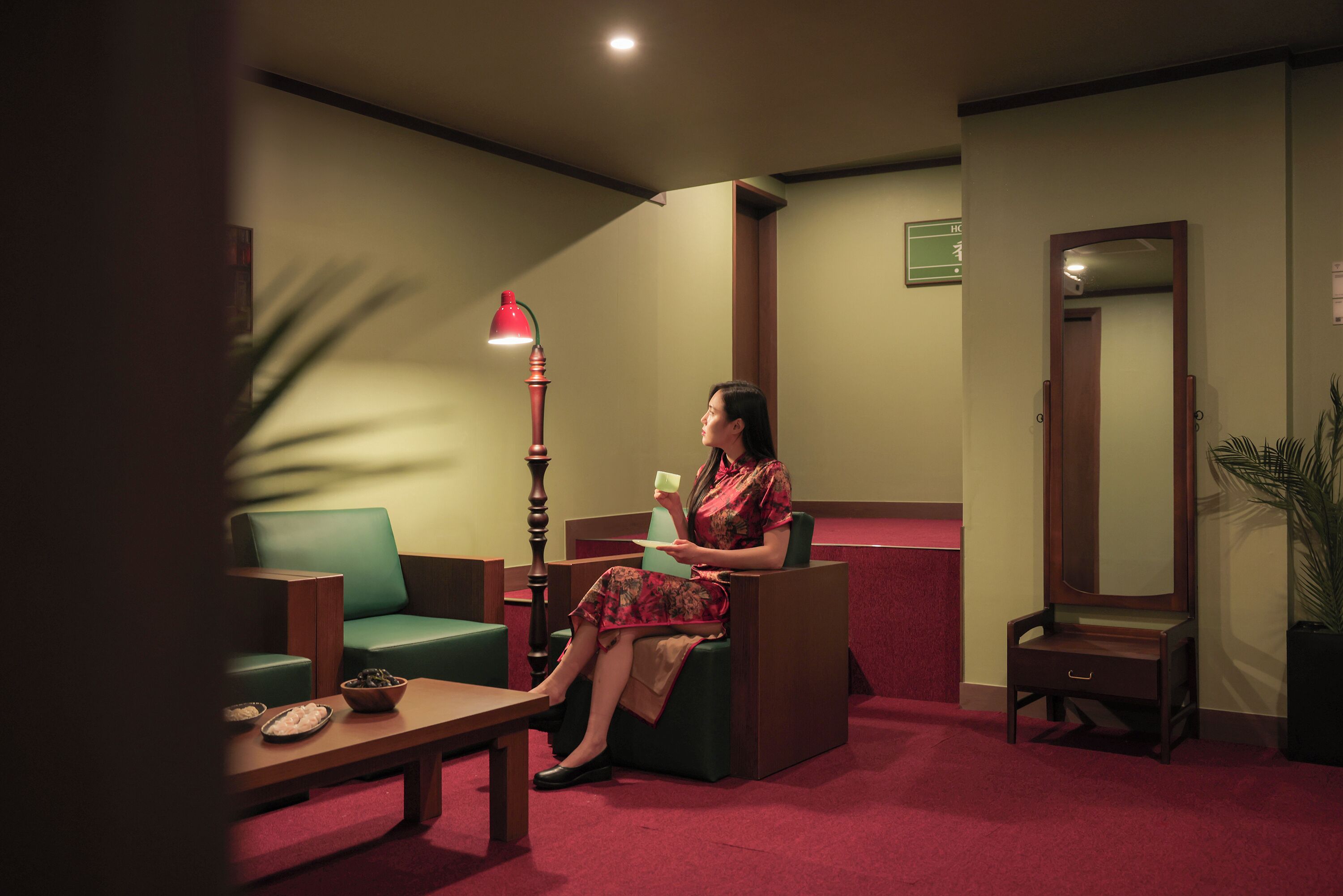 A woman seated in a room with a green sofa, a wooden table, and a red lamp.