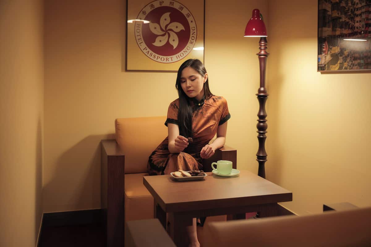 A woman sitting at a wooden table with a green cup and a plate of cookies. There is a red lamp and a framed sign on the wall behind her.